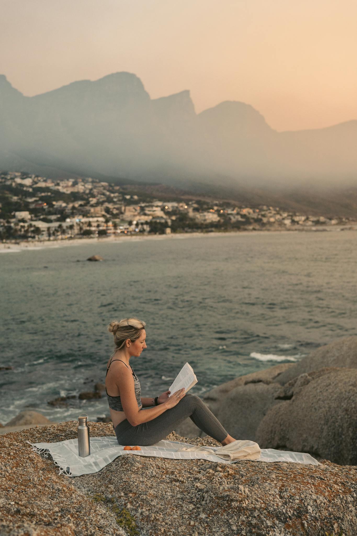 A woman enjoys reading by the ocean with a mountain view in Cape Town during sunset.